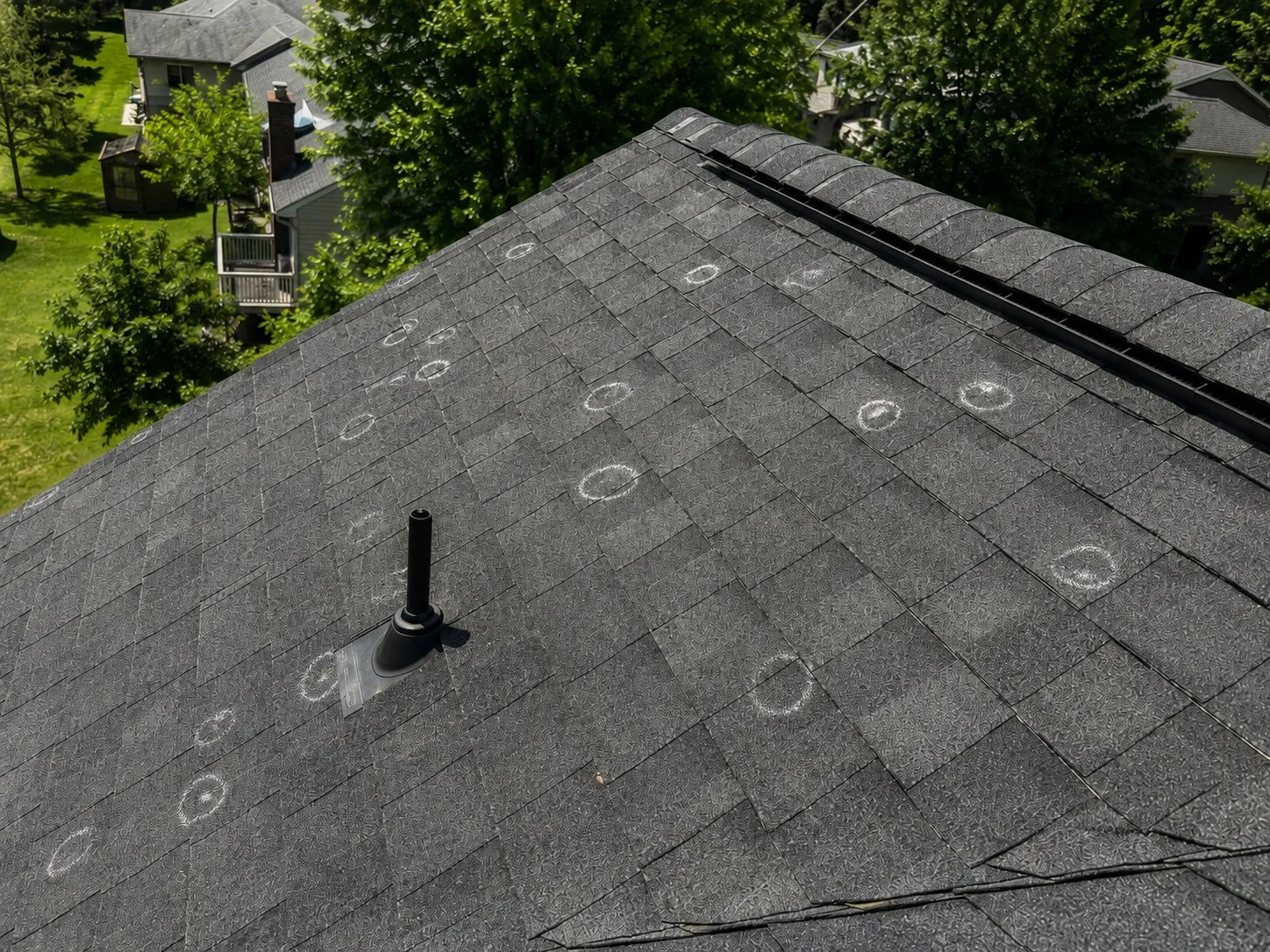 A dark asphalt shingle roof photographed from above, with hail impact bruises circled in white chalk across the slope and around the vent boot.
