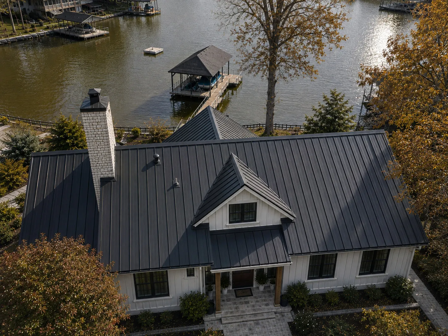 A white board-and-batten lakefront home seen from above with a new dark standing seam metal roof, its long panel runs unbroken from ridge to eave, the covered dock and open water behind it.